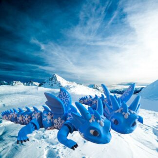 Two articulated 3D-printed blue ice dragons with white snowflake patterns on their scales, posed on a snowy mountain landscape under a blue sky